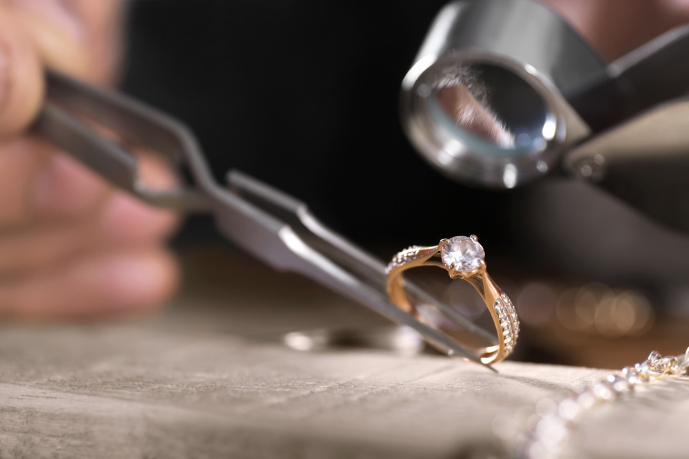 A close up of a jeweler using a magnifying glass and tweezers to hold and inspect a diamond ring