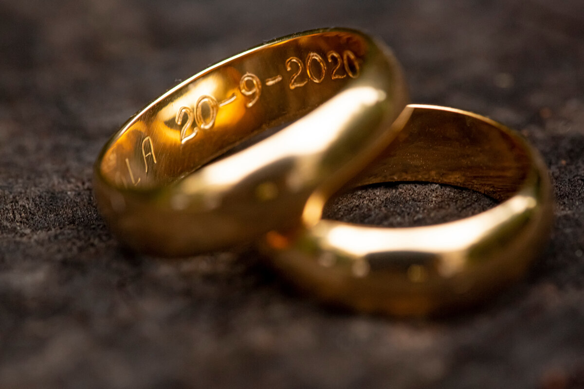 A pair of yellow gold men's wedding bands sit against a stonr background. On the inside of one band, a date is engraved. 